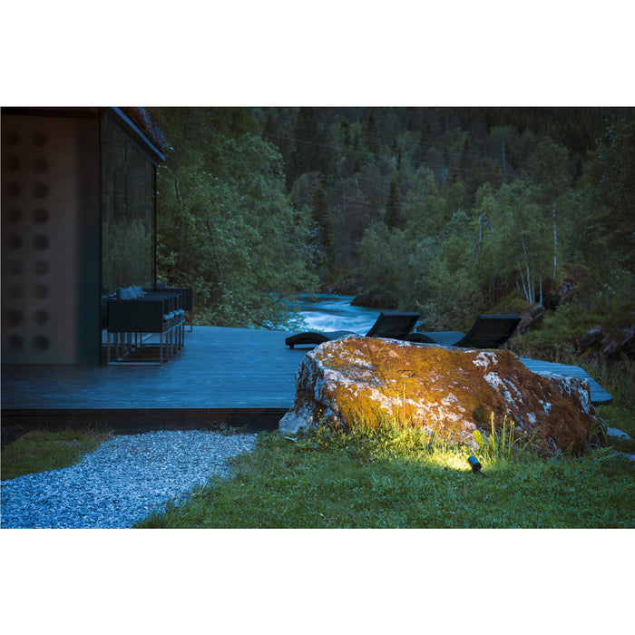 Black outdoor spotlight illuminating mossy rock near a wooden deck and river.