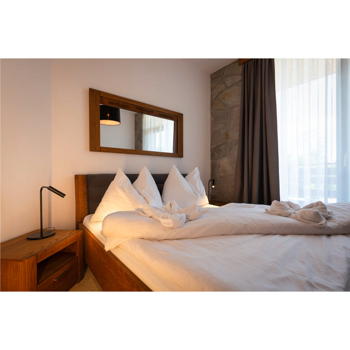 Bedroom with wood bed frame, white linens, wood nightstand, and wood-framed mirror.