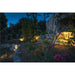 Bronze outdoor spotlight illuminating a stone retaining wall and garden at dusk.