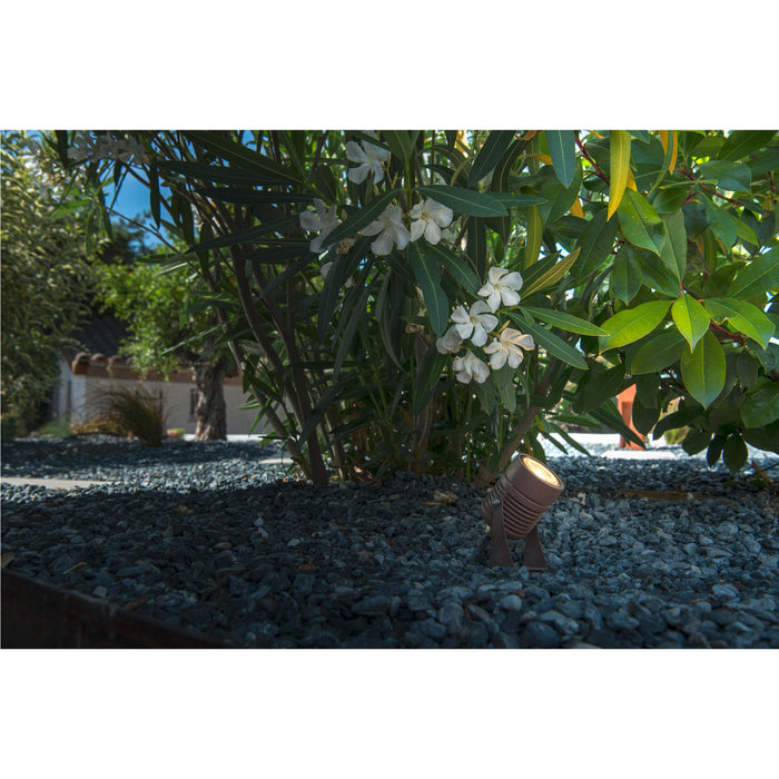 Bronze finish landscape spotlight, ground mounted among gray gravel and greenery.