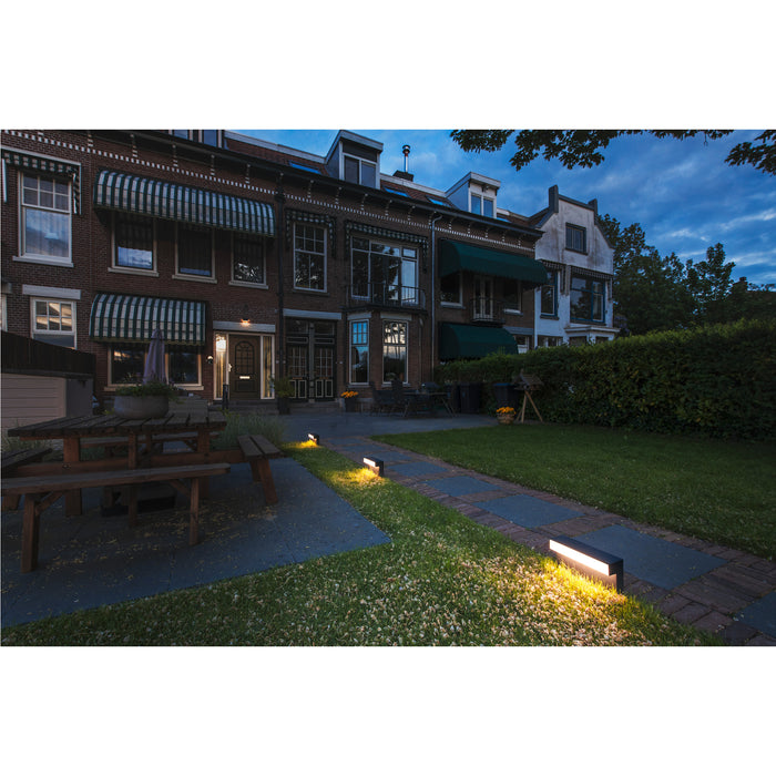 Black rectangular path light illuminating a stone and grass walkway in front of brick buildings.
