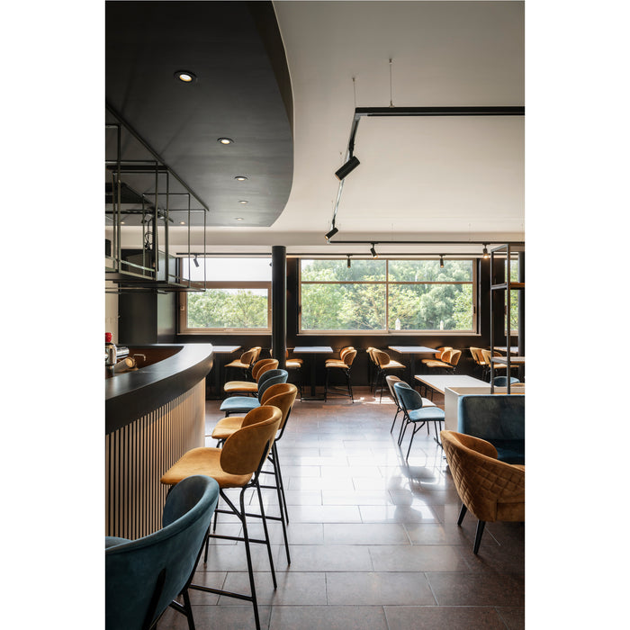 Restaurant interior with velvet upholstered chairs in blue and gold, black metal frames, and wood bar.