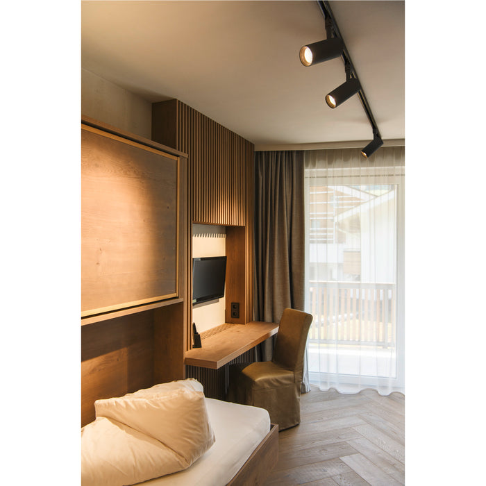 Modern bedroom with wood Murphy bed, desk, leather chair, herringbone floor, and black track lighting.