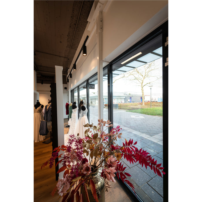 Bridal shop interior with wedding dresses on mannequins, floral arrangement in metallic vase, and black framed windows.