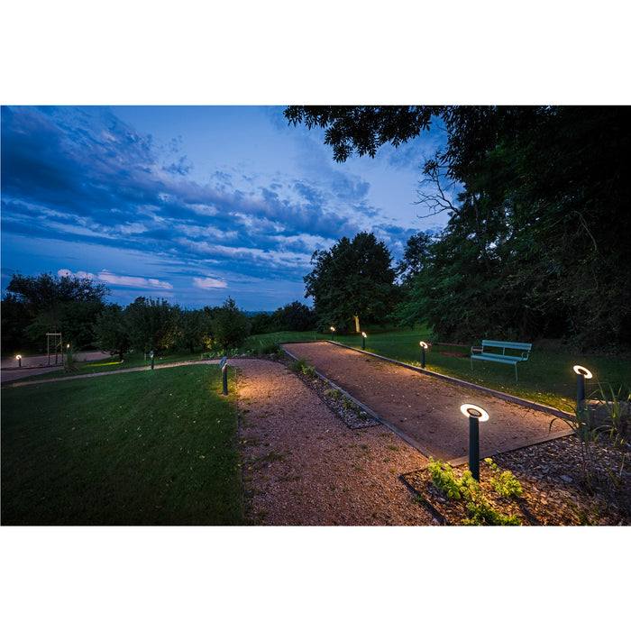 Black metal bollard lights with round diffuser, ground-mounted, illuminating a bocce court at dusk.