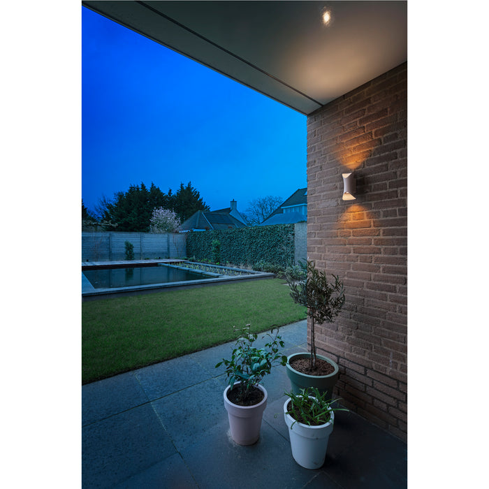 White wall-mounted sconce lighting a brick wall on a patio overlooking a garden with a pond.