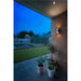 White wall-mounted sconce lighting a brick wall on a patio overlooking a garden with a pond.