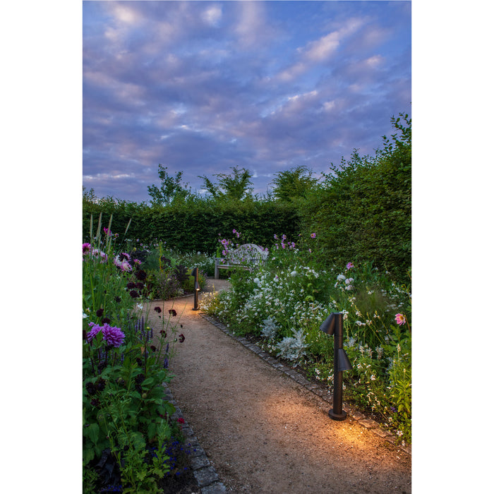 Black metal path lights illuminating a garden path with flowers and a bench.