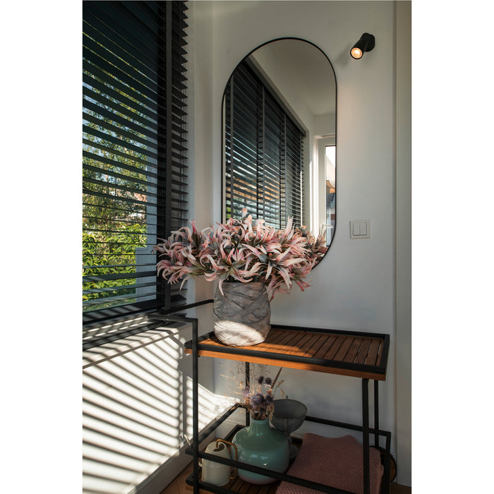 Black framed oval mirror above wood and metal console table with gray vase and pink flowers.