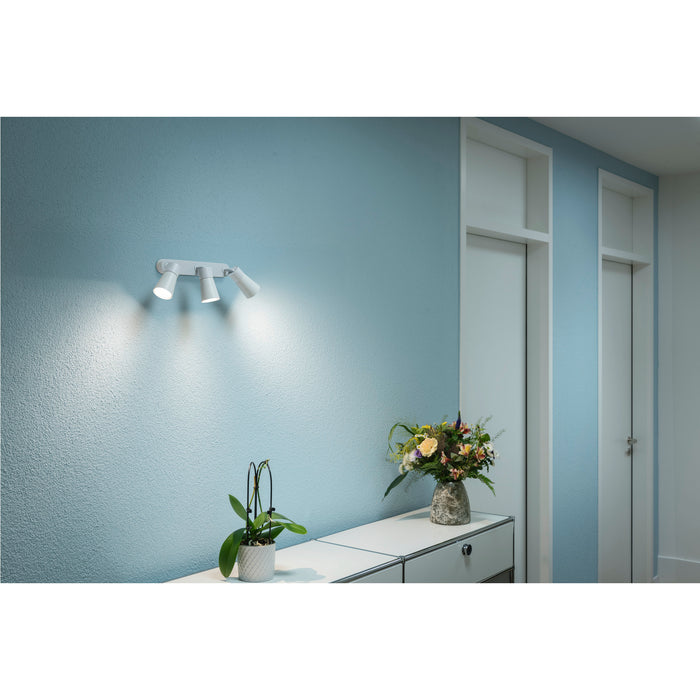 White metal wall-mounted spotlight with three adjustable heads, illuminating a blue textured wall.