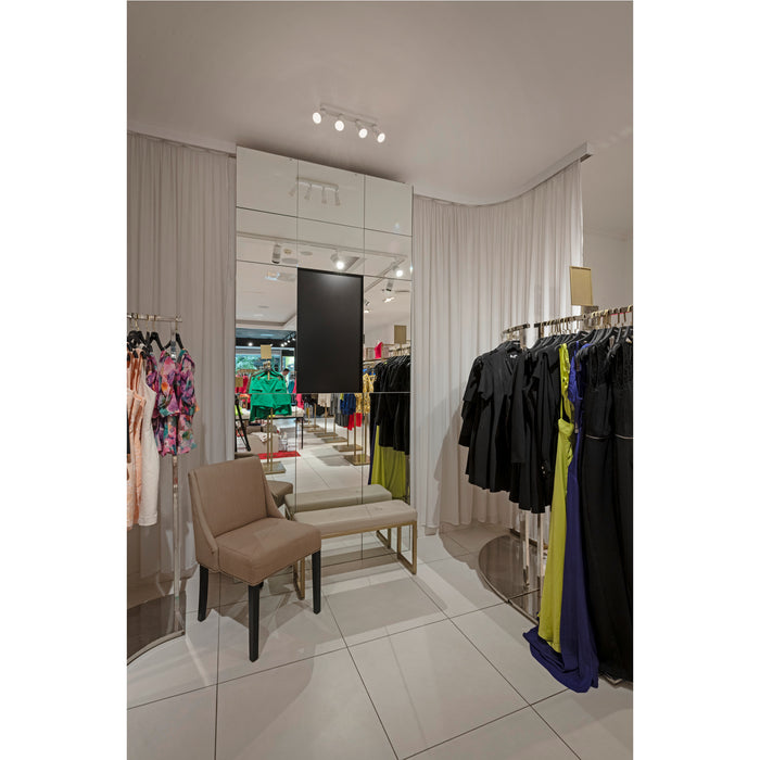Clothing store fitting room with beige upholstered chair and bench, mirrored wall, and clothing racks.