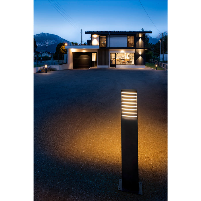 Black bollard light fixture, metal, with horizontal light bars, ground mounted, illuminating a driveway at dusk.