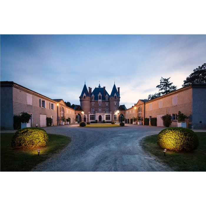 Brick chateau with blue roof and turrets, gravel driveway, and symmetrical outbuildings at dusk.