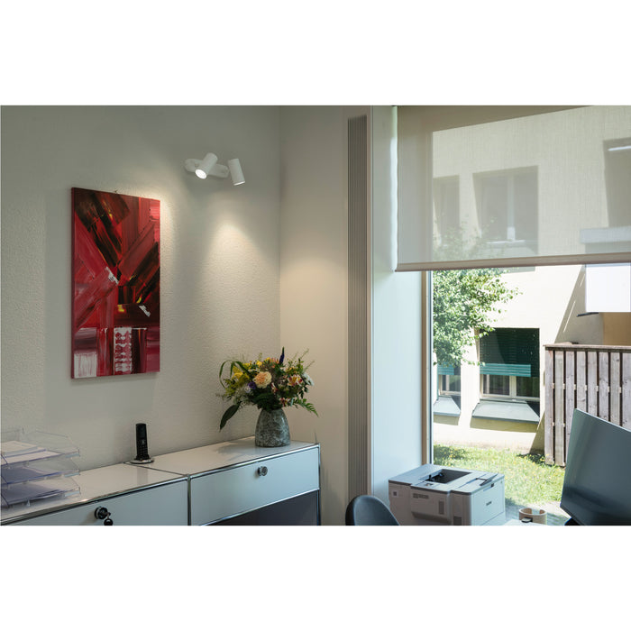 White wall-mounted spotlight with two adjustable heads, illuminating a painting in an office setting.