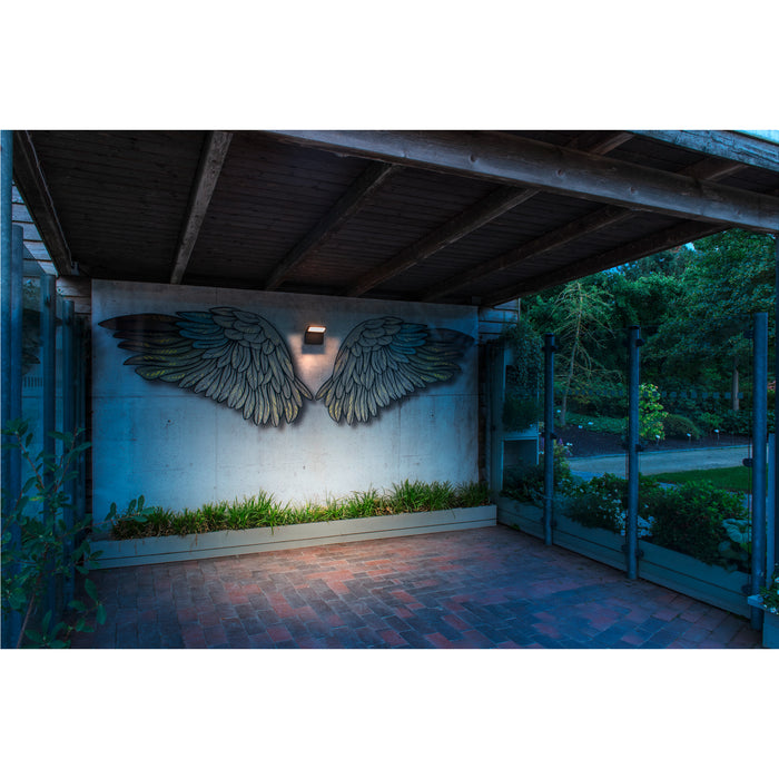 Wall-mounted metal angel wings art, gray finish, illuminated by a sconce.