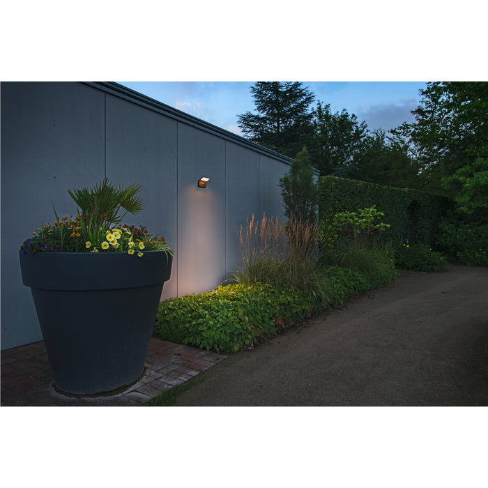 Dark gray wall-mounted outdoor light illuminating a large dark gray planter with flowers in a garden setting.