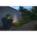 Dark gray wall-mounted outdoor light illuminating a large dark gray planter with flowers in a garden setting.