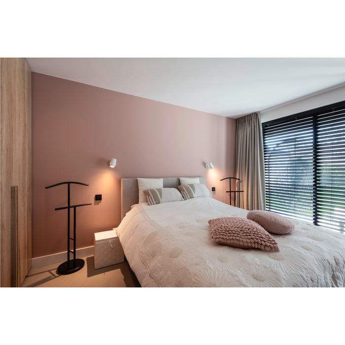 Bedroom with white floral-patterned quilt, pink pillows, black metal valet stand, and wall-mounted white sconces.