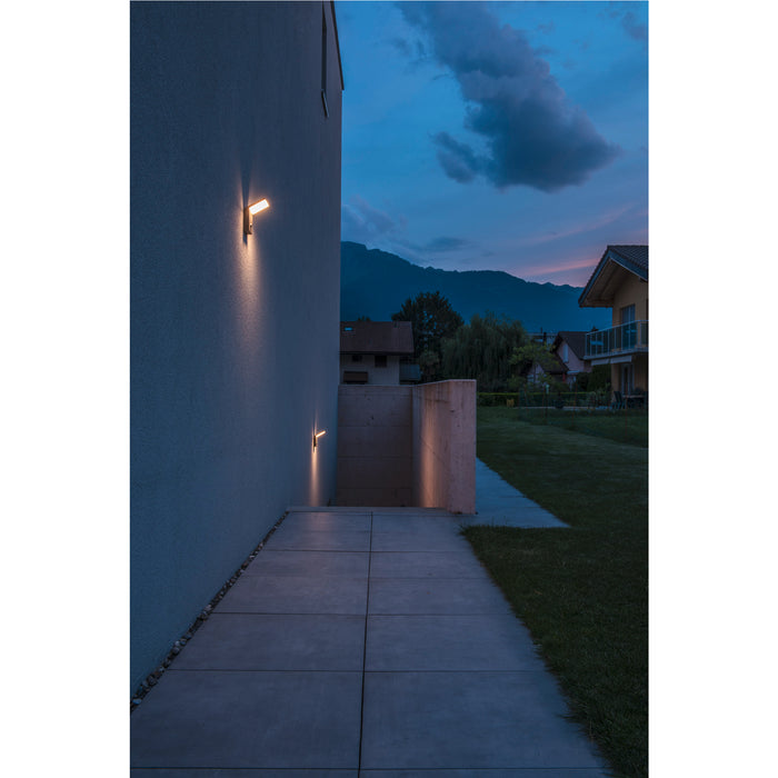 Modern gray wall-mounted LED light fixture illuminating a walkway at dusk.